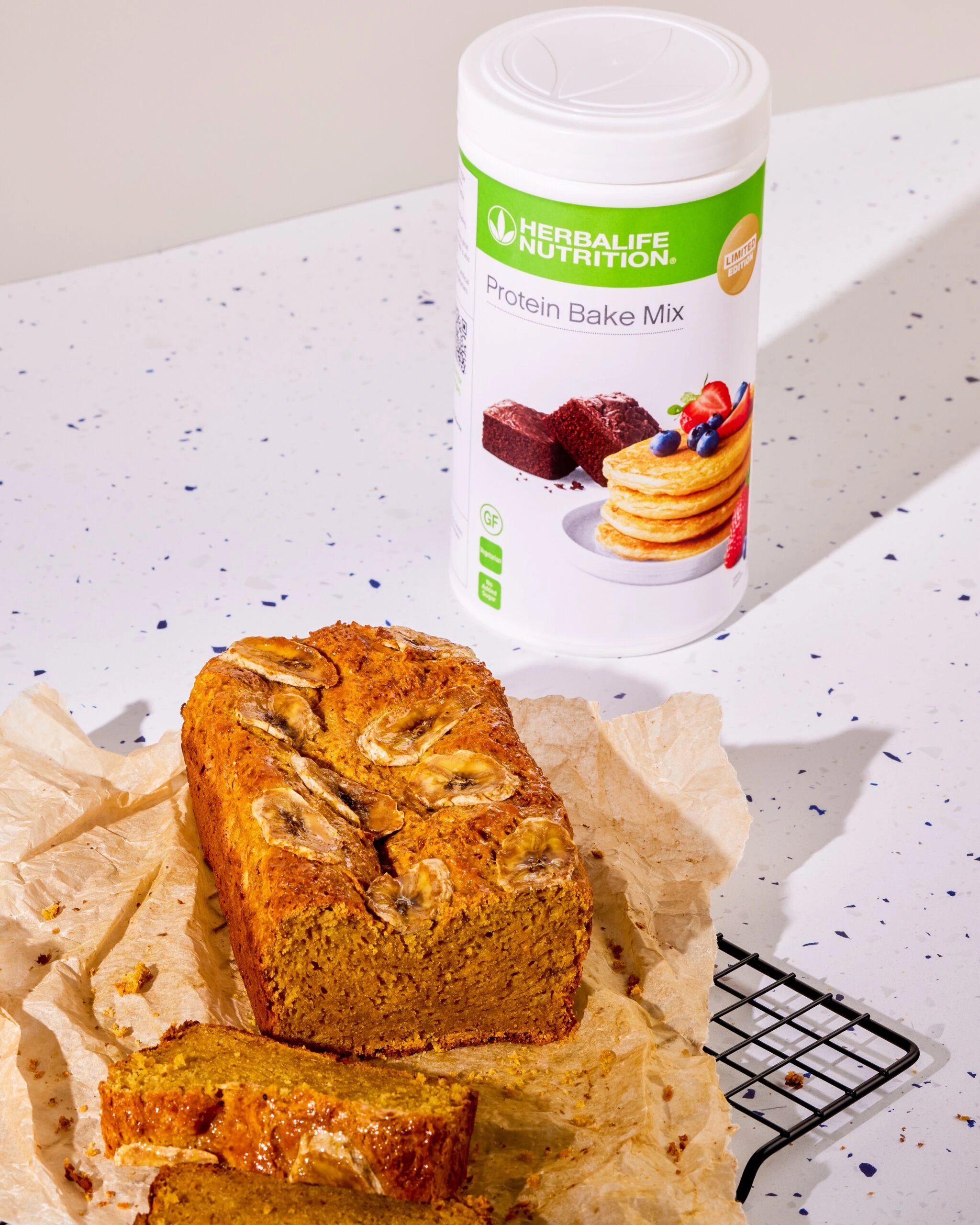 Herbalife Protein Bake Mix ile Hazırlanmış Fit Muzlu Kek Herbalife Protein Bake Mix kutusu ve yanında fırından yeni çıkmış, pişirme kağıdı üzerinde dilimlenmiş yüksek proteinli fit muzlu kek (banana bread).